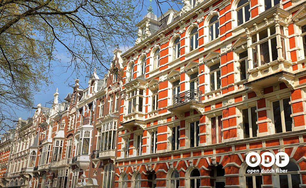 Row of ornate red and white brick buildings with large windows under a clear blue sky; tree branches in the foreground.