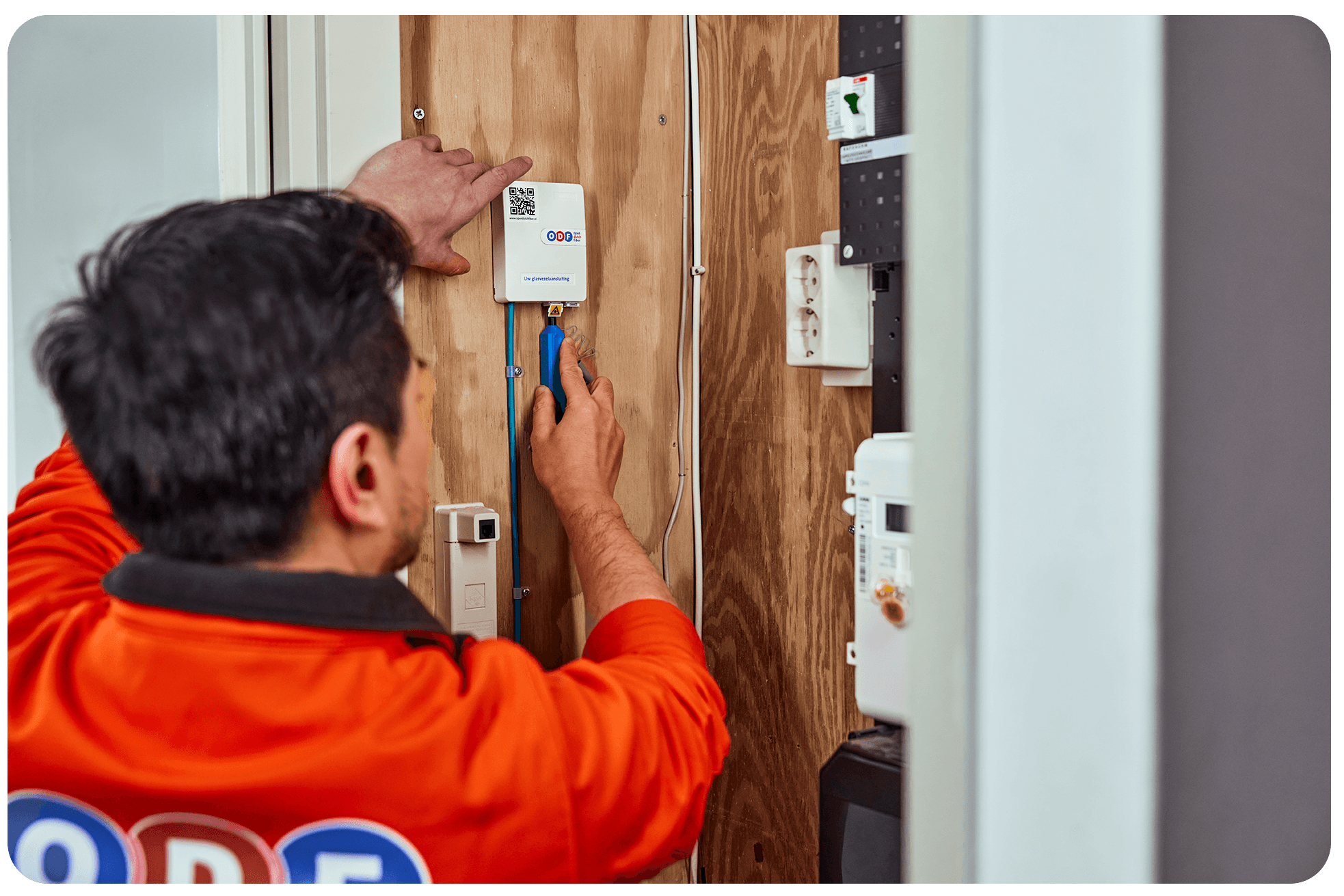 Technician in an orange ODF branded shirt installing a white communication or internet box (with QR code) onto a wooden mounting board, likely performing a fiber optic installation in a utility closet or electrical panel area.